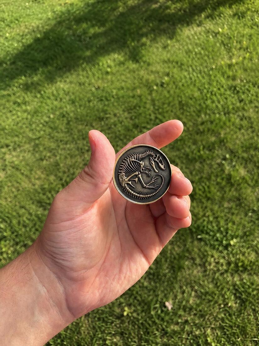 Antiqued brass coin with a raised dinosaur skeleton relief, held in a hand