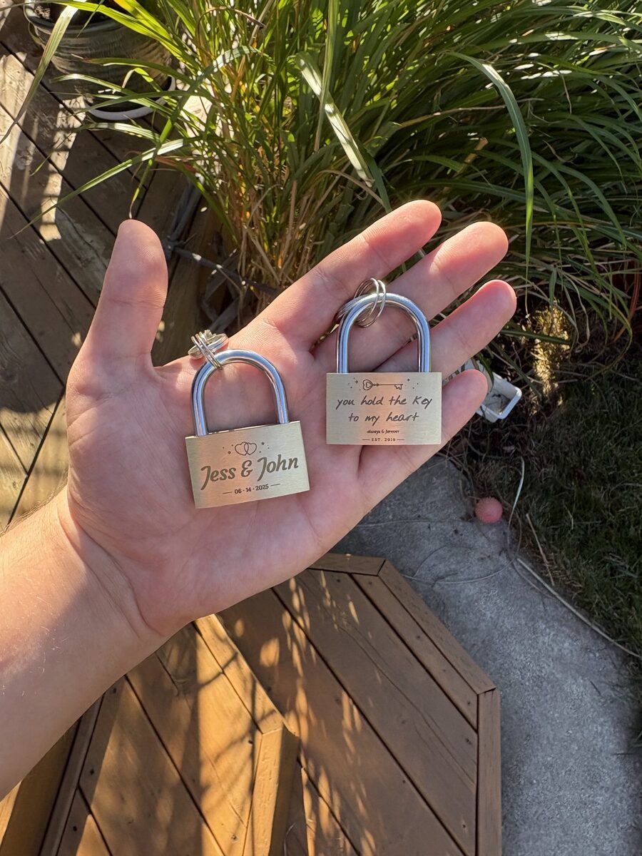 Two brass padlocks held in an open palm, one engraved Jess & John 06·14·2025 beneath interlocking hearts, the other engraved you hold the key to my heart, always & forever, est. 2019